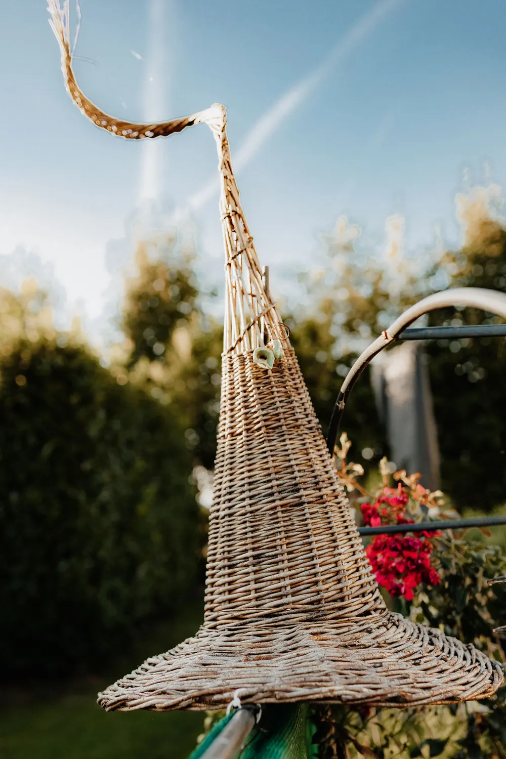 Ein geflochtener Hexenhut aus Weide hängt im Garten, dahinter rote Blüten und Bäume