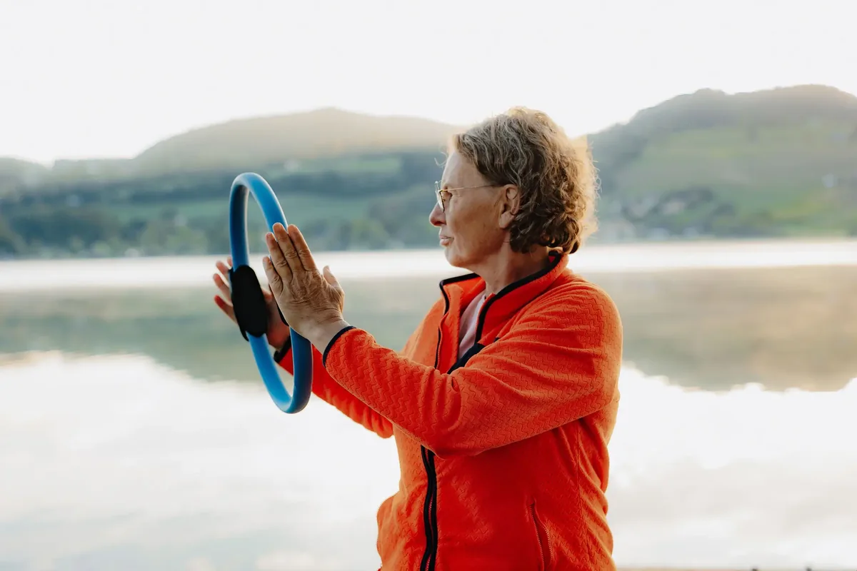 Renate Troppmair hält einen Pilates-Ring in beiden Händen, im Hintergrund ein nebliger See