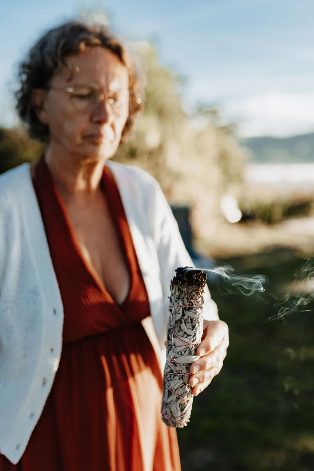 Renate Troppmair hält ein rauchendes Salbeibündel in der Hand, hinter ihr der See im Morgenlicht
